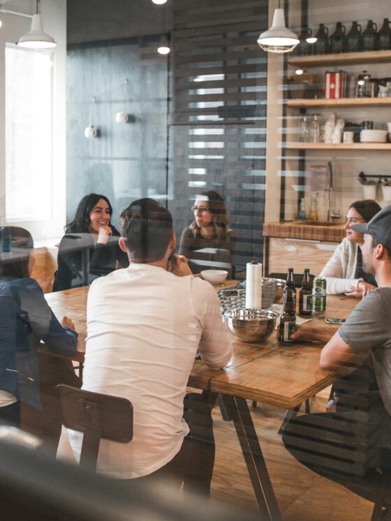 Group of people seated around a wooden table in a modern cafe, chatting and sharing drinks in a casual meeting.
