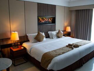 Hotel room with a large bed, two beige throw pillows, wooden headboard wall, and warm bedside lamps on each side.