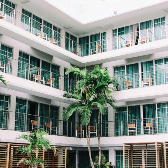 White multi-story apartment building with glass doors, balconies, and wooden chairs; tropical palms in front.