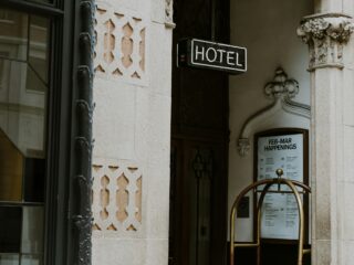 Hotel entrance with ornate stone archway and a neon 'HOTEL' sign above the door, a luggage cart nearby.