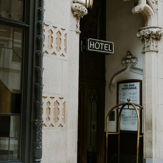 Hotel entrance with ornate stone archway and a neon 'HOTEL' sign above the door, a luggage cart nearby.