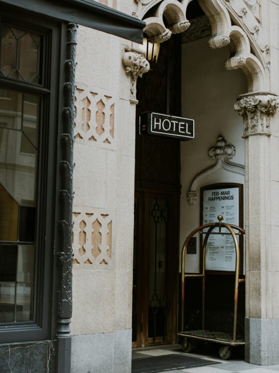 Hotel entrance with ornate stone archway and a neon 'HOTEL' sign above the door, a luggage cart nearby.