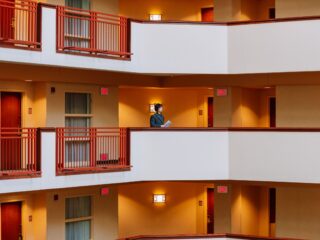 Person in a gray blazer standing on a hotel atrium walkway, holding papers amid orange walls and red railings.