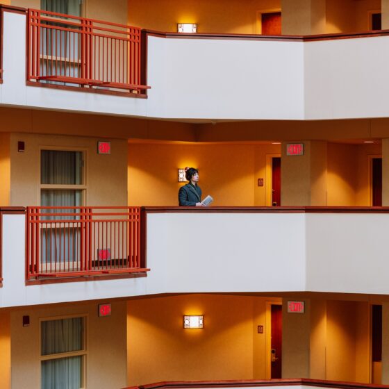 Person in a gray blazer standing on a hotel atrium walkway, holding papers amid orange walls and red railings.