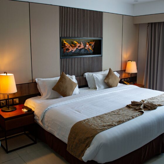Hotel room with two large beds, neutral beige wall panels, and decorative wood slats behind a built‑in art feature; lamps on bedside tables, and closed curtains to the right.