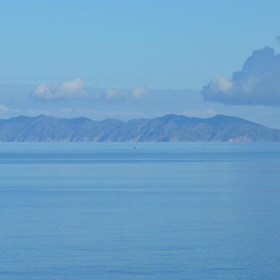 Calm blue sea with a distant mountainous island on the horizon under a pale cloud-filled sky.