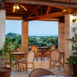 Outdoor dining terrace with beige wicker chairs and round tables, stone columns, and warm lighting, overlooking a landscape.