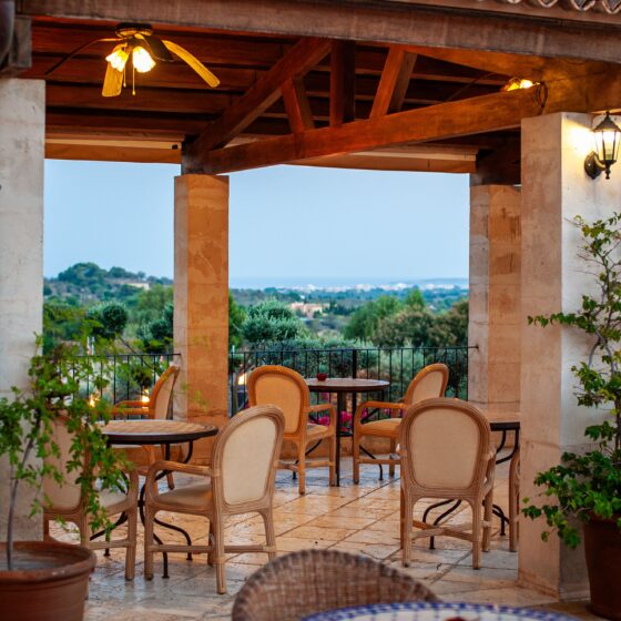 Outdoor dining terrace with beige wicker chairs and round tables, stone columns, and warm lighting, overlooking a landscape.
