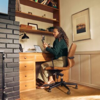Woman sits at a wooden desk in a cozy home office, looking at a phone with a laptop open beside her, brick fireplace on the left and shelves above.