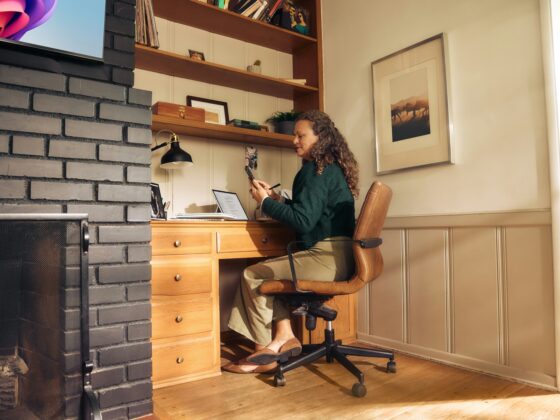 Woman sits at a wooden desk in a cozy home office, looking at a phone with a laptop open beside her, brick fireplace on the left and shelves above.