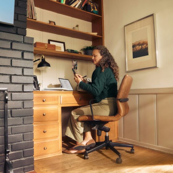 Woman sits at a wooden desk in a cozy home office, looking at a phone with a laptop open beside her, brick fireplace on the left and shelves above.