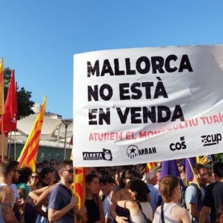 Crowd of protesters holding a large banner that reads 'Mallorca no está en venta' with yellow-red flags on a sunny day.