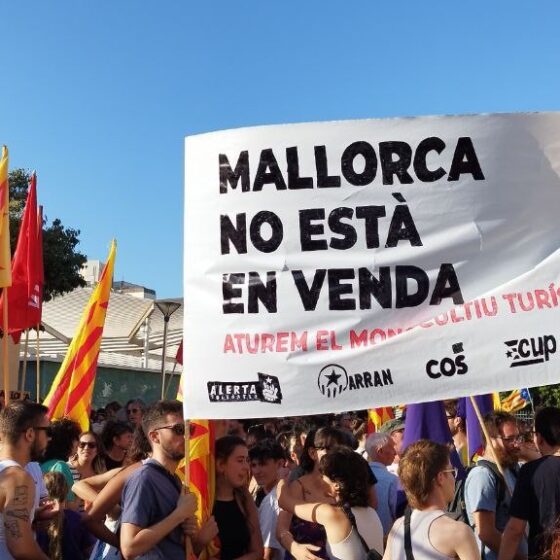 Crowd of protesters holding a large banner that reads 'Mallorca no está en venta' with yellow-red flags on a sunny day.