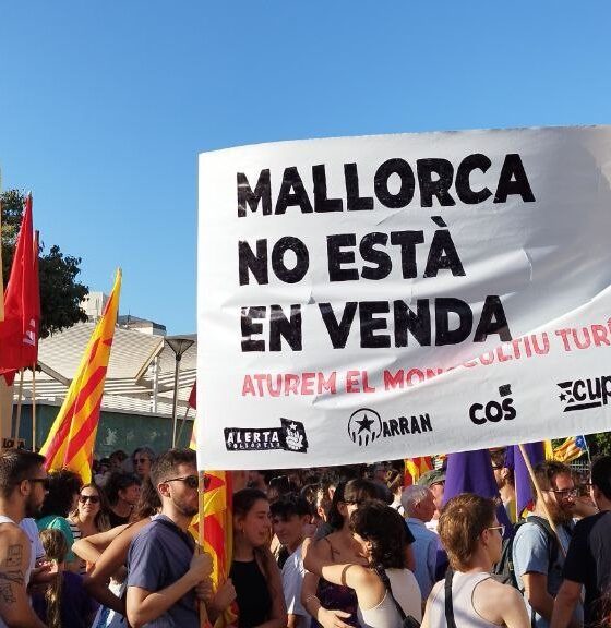 Crowd of protesters holding a large banner that reads 'Mallorca no está en venta' with yellow-red flags on a sunny day.