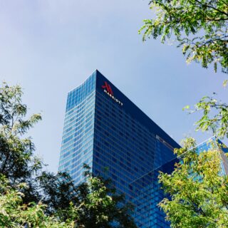 Blue glass skyscraper with Marriott logo, partially visible behind green tree branches against a clear blue sky.