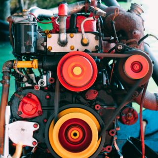 Close-up of a vintage engine showing red components, belts, and bright orange/yellow pulleys in the foreground with a rusted housing in the background.