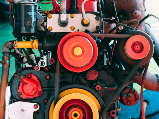 Close-up of a vintage engine showing red components, belts, and bright orange/yellow pulleys in the foreground with a rusted housing in the background.