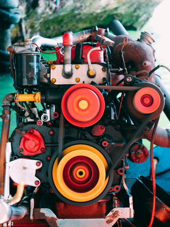 Close-up of a vintage engine showing red components, belts, and bright orange/yellow pulleys in the foreground with a rusted housing in the background.