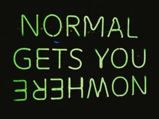 Neon green uppercase sign spelling out words (appears to read) 'NORMAL GETS YOU EMPOWERMENT' against a dark background, viewed from below.