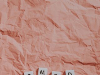 Crinkled pink paper background with white Scrabble tiles spelling 'PMDD' (letters P, M, D, D) laid near the bottom edge, each tile showing a small game score.