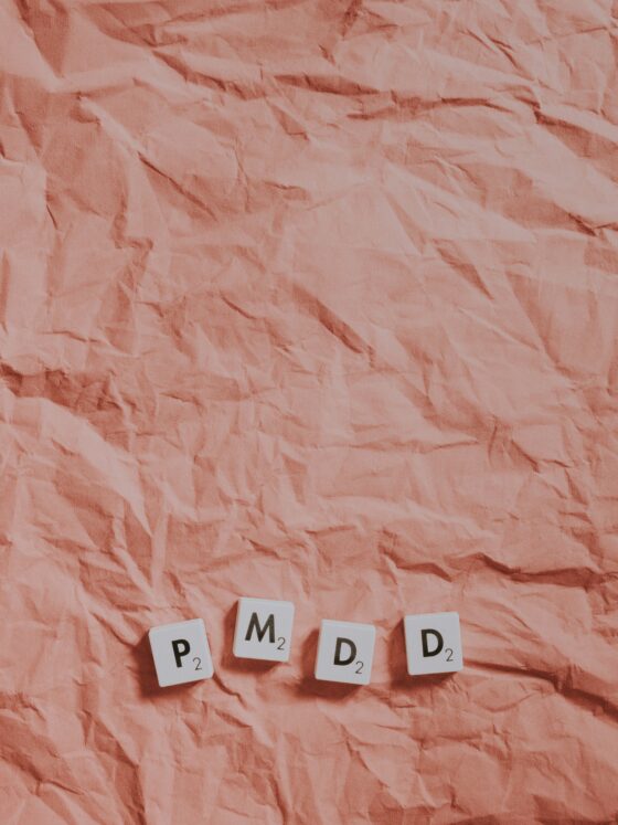 Crinkled pink paper background with white Scrabble tiles spelling 'PMDD' (letters P, M, D, D) laid near the bottom edge, each tile showing a small game score.