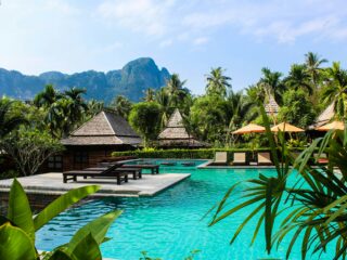 Resort pool area with turquoise water, thatched-roof cabanas, lounge chairs, and lush tropical palms under a blue sky.