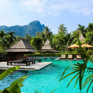 Resort pool area with turquoise water, thatched-roof cabanas, lounge chairs, and lush tropical palms under a blue sky.