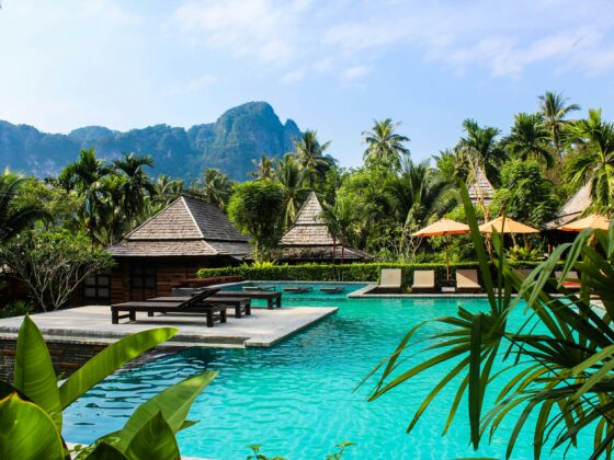 Resort pool area with turquoise water, thatched-roof cabanas, lounge chairs, and lush tropical palms under a blue sky.
