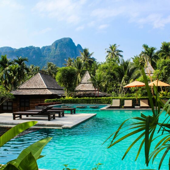 Resort pool area with turquoise water, thatched-roof cabanas, lounge chairs, and lush tropical palms under a blue sky.