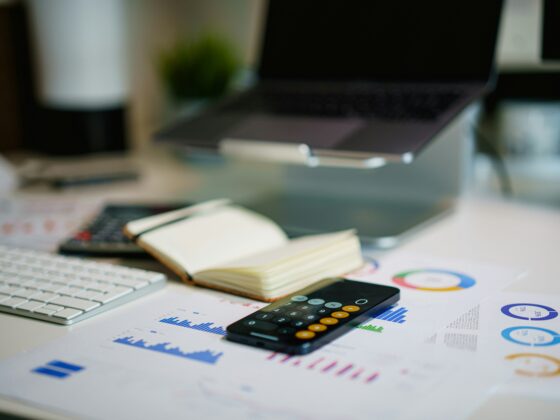 Office desk with a laptop, external keyboard, calculator, open notebook, and printed charts.