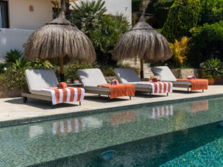 Poolside scene with four lounge chairs under thatched umbrellas and orange towels along a sunny pool edge