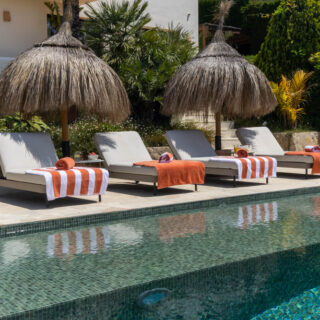 Poolside scene with four lounge chairs under thatched umbrellas and orange towels along a sunny pool edge