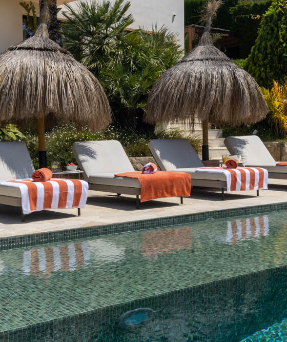 Poolside scene with four lounge chairs under thatched umbrellas and orange towels along a sunny pool edge