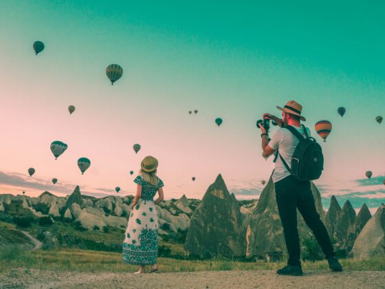 Man with a backpack and camera photographs a field of colorful hot air balloons against a clear sky, with a woman in a patterned dress nearby on rocky terrain.