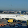 White Vueling Airlines Airbus A320 at a gate, yellow engine nacelles, jet bridge attached, ground crew and vehicles nearby