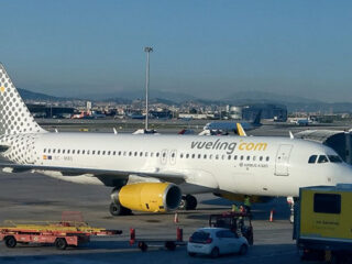 White Vueling Airlines Airbus A320 at a gate, yellow engine nacelles, jet bridge attached, ground crew and vehicles nearby