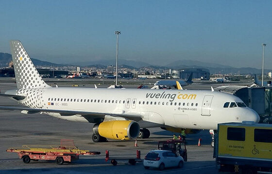 White Vueling Airlines Airbus A320 at a gate, yellow engine nacelles, jet bridge attached, ground crew and vehicles nearby
