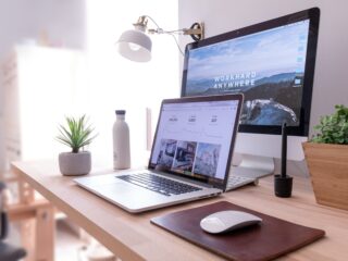 Home office desk with a laptop showing analytics, a second monitor displaying a mountain wallpaper, a desk lamp, plant, water bottle, and mouse on a wooden desk.