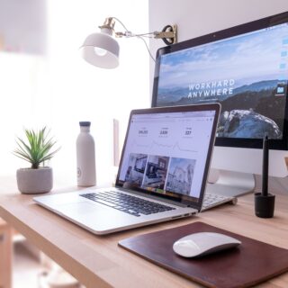 Home office desk with a laptop showing analytics, a second monitor displaying a mountain wallpaper, a desk lamp, plant, water bottle, and mouse on a wooden desk.