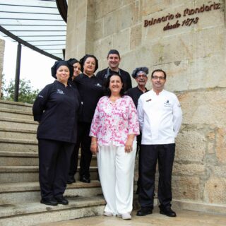 Group of chefs and staff posing on stone stairs outside a building with a carved stone wall and sign above them (Balneario de Mondariz).