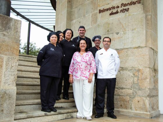 Group of chefs and staff posing on stone stairs outside a building with a carved stone wall and sign above them (Balneario de Mondariz).
