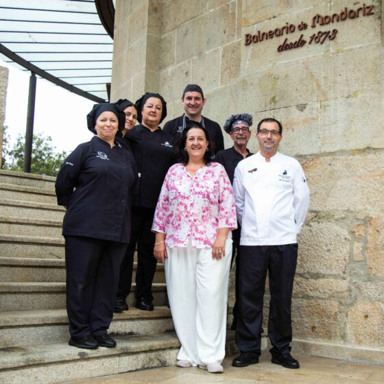 Group of chefs and staff posing on stone stairs outside a building with a carved stone wall and sign above them (Balneario de Mondariz).