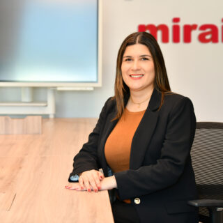 Professional woman in a blazer sits at a wooden conference table, smiling, with a red 'mirai' logo on the wall behind her.