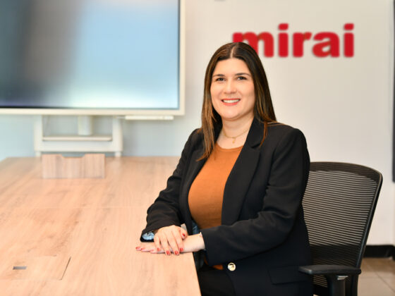 Professional woman in a blazer sits at a wooden conference table, smiling, with a red 'mirai' logo on the wall behind her.