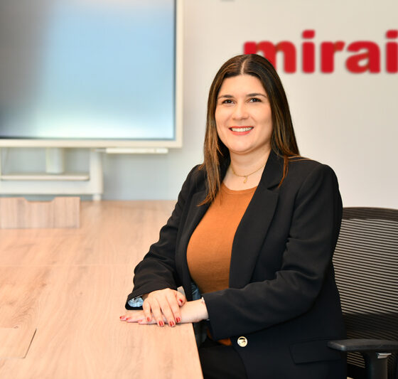 Professional woman in a blazer sits at a wooden conference table, smiling, with a red 'mirai' logo on the wall behind her.
