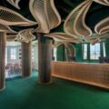 Interior of a modern lobby/bar with green walls, sculptural wooden slat columns, and a curved wooden reception desk.
