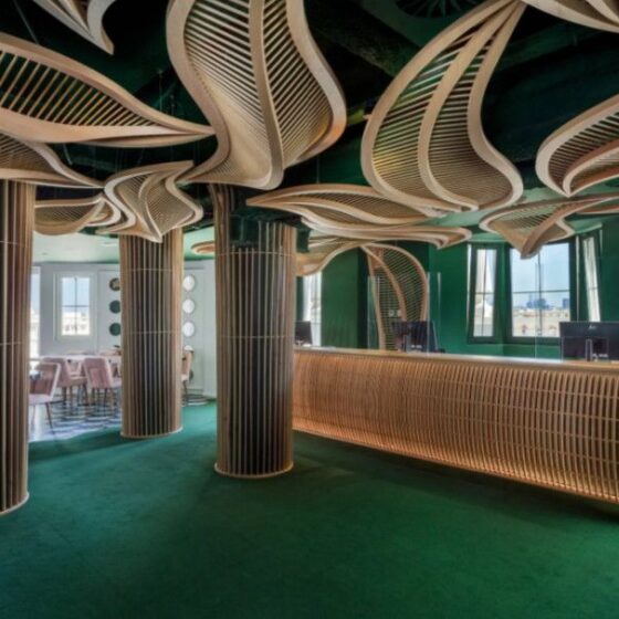 Interior of a modern lobby/bar with green walls, sculptural wooden slat columns, and a curved wooden reception desk.
