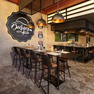 Modern burger shop interior with a long communal table, black stools, industrial pendant lights, and vintage Route 66 signs on a concrete wall; warm, inviting atmosphere.