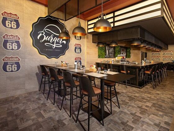 Modern burger shop interior with a long communal table, black stools, industrial pendant lights, and vintage Route 66 signs on a concrete wall; warm, inviting atmosphere.