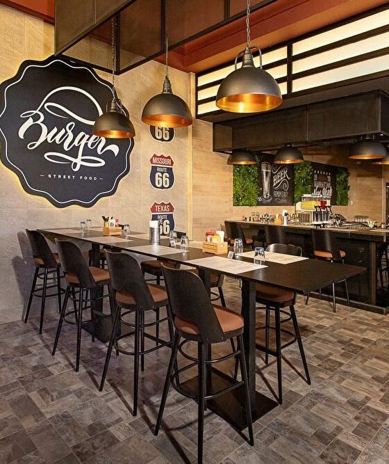 Modern burger shop interior with a long communal table, black stools, industrial pendant lights, and vintage Route 66 signs on a concrete wall; warm, inviting atmosphere.